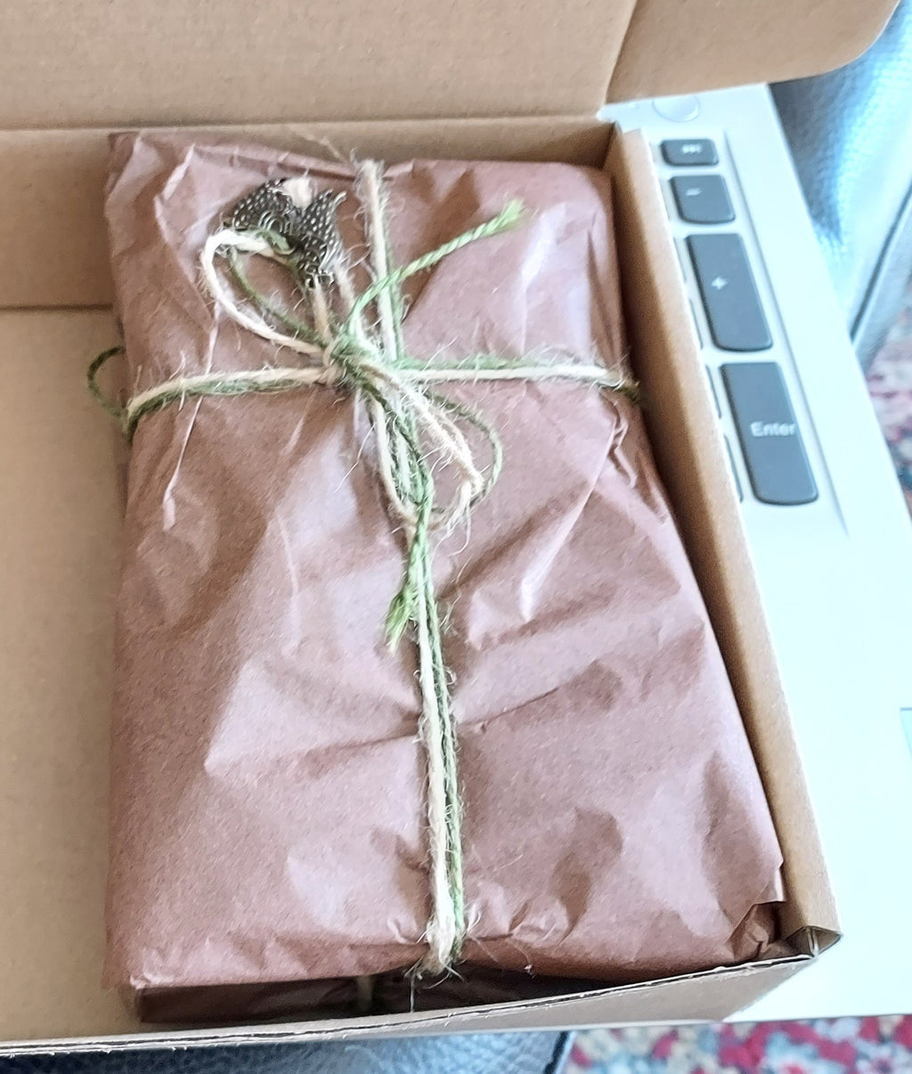 A brown kraft paper wrapped package tied with twine and a small decorative sprig, sitting inside an open cardboard box next to a laptop keyboard.